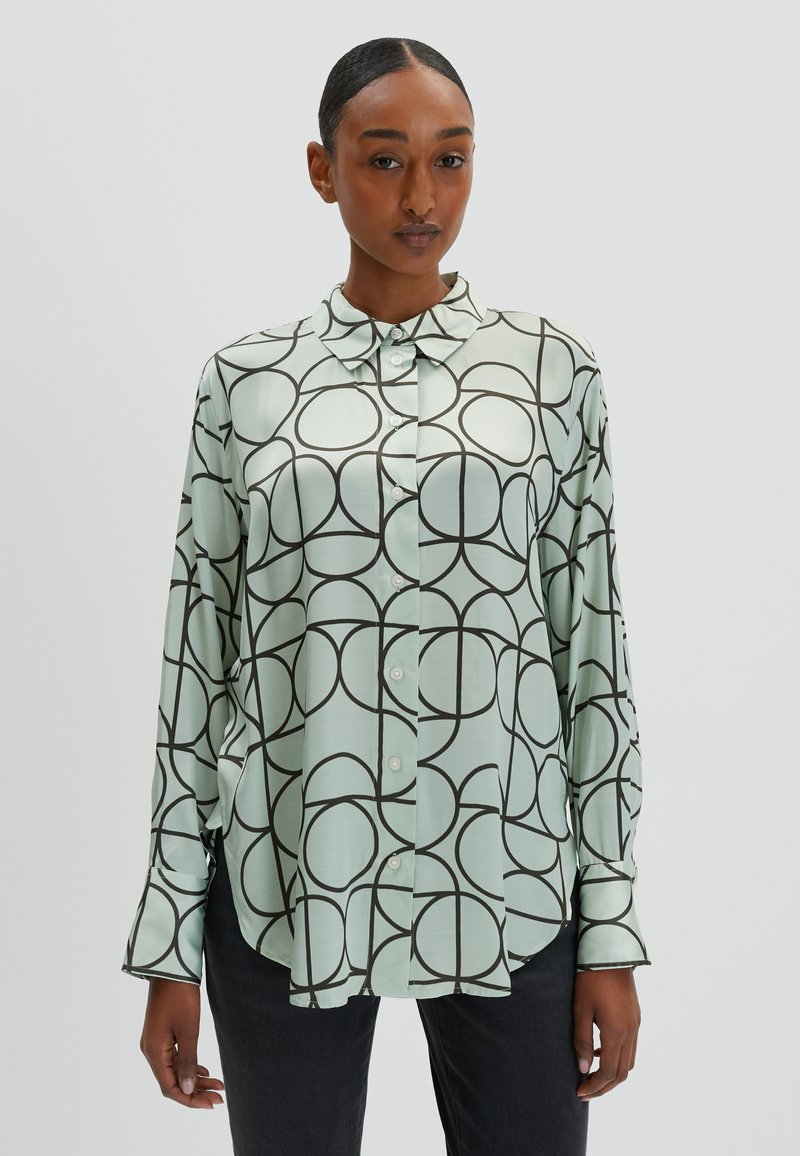 Mintgroene blouse met een geometrisch patroon van overlappende zwarte cirkels, knoopsluiting aan de voorkant en lange mouwen met manchet.