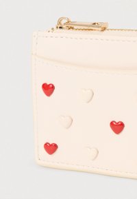 Porte-cartes couleur crème orné de petites décorations en forme de cœur rouge et blanc brillantes. Fermeture sécurisée grâce à une fermeture éclair dorée en haut.
