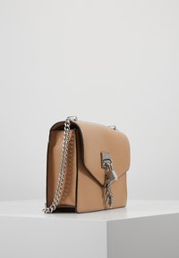 Beżowa skórzana torba na ramię z srebrnym łańcuchem i dekoracyjnym srebrnym zamkiem oraz zawieszką, stojąca na białym podium wystawowym.