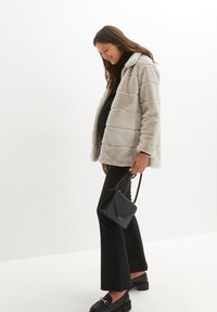 Giacca imbottita in pelliccia sintetica, di un grigio chiaro, con cuciture orizzontali, tasche frontali e una vestibilità comoda. Indossata sopra una maglietta nera e pantaloni a palazzo.