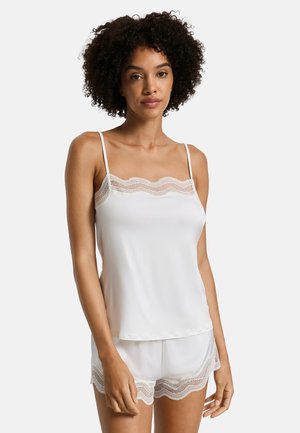 Donna con capelli ricci che indossa una canottiera bianca con pizzo e pantaloncini coordinati, in piedi davanti a uno sfondo chiaro e uniforme.