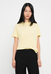 Żółta koszulka z krótkim rękawem, okrągłym dekoltem i białym nadrukiem logo "TOMMY HILFIGER EST. 1985", połączona z czarnymi spodniami.