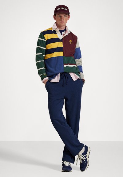 Ungdommelig mann iført en flerfarget striped rugby genser, marineblå joggebukser, blå sneakers og en maroon baseball caps med teksten "NEW ENGLAND".