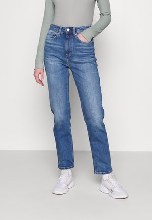 Personne portant un jean bleu taille haute, un haut vert clair à manches longues, et des baskets blanches, debout avec une main dans la poche.