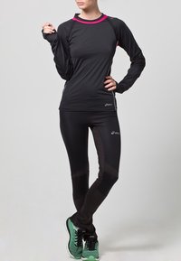Persona che indossa una maglietta a maniche lunghe nera Asics e leggings con dettagli rosa, scarpe da corsa verdi, in piedi con una mano sul fianco.