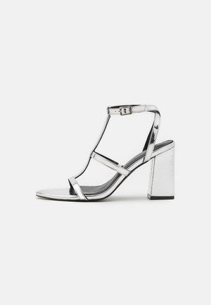 Sandalie metalice argintii cu toc, cu un design în T, cataramă ajustabilă pentru glezna și toc pătrat, având o suprafață netedă și reflexivă.