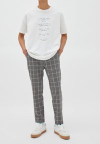 Persona in piedi con le mani in tasca, indossa una maglietta bianca con testo, pantaloni grigi a quadri e sneakers bianche con suola in gomma, su uno sfondo bianco uniforme.