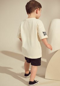 Creme kortærmet skjorte med en glat tekstur, sorte shorts og sorte sneakers. Skjorten har et "Mickey Mouse"-grafik på ærmet.