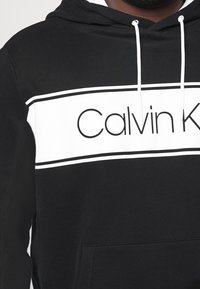 Felpa nera con accenti bianchi, dotata di un logo "Calvin Klein" centrato. Realizzata in materiale morbido, con una tasca frontale e cappuccio con cordino.