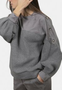 Donna che indossa una felpa grigia bicolore con bottoni sulla spalla e pantaloni grigi abbinati, con una mano che tocca i capelli e l'altra in tasca.