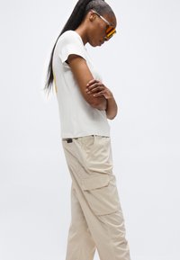 Γυναίκα με μακριά μαλλιά φοράει λευκό μπλουζάκι, μπεζ παντελόνι cargo και αντανακλαστικά πορτοκαλί γυαλιά ηλίου, στέκεται με σταυρωμένα τα χέρια μπροστά σε λευκό φόντο.