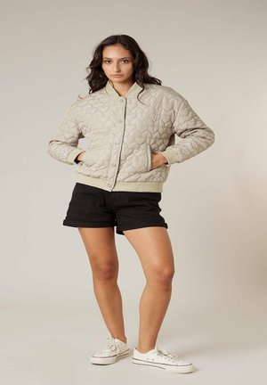 Femme portant une veste matelassée beige à motifs de cœurs, un short noir et des baskets blanches, debout les mains dans les poches sur un fond uni.