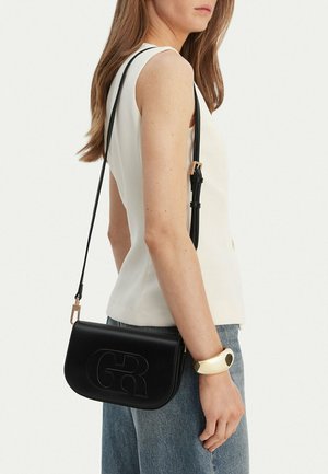 Mujer con top blanco sin mangas y jeans azules que lleva un bolso negro de cuero con logo "GR" en relieve y una pulsera dorada en la muñeca.