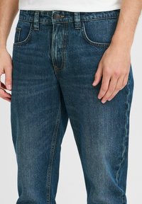 Dunkelblaue Denim-Jeans im klassischen Schnitt mit genähten Akzenten und einem Knopfverschluss an der Taille. Der Stoff zeigt leichte Verblassung.