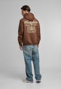 Brun hettegenser med frontlomme og hvit trykt design på ryggen; løse lyseblå jeans; nøytrale fargede joggesko.