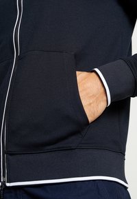 Marinblå zip-up-jacka med en slät textur, vita detaljer på muddar och dragkedja. Har framfickor och en åtsittande passform.