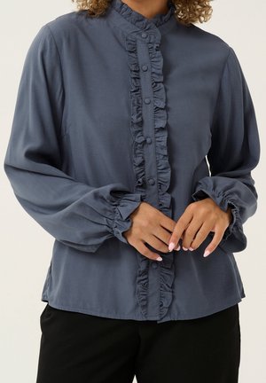 Kobieta ubrana w ciemnoniebieską bluzkę z długim rękawem, z falbankową plisą na guziki oraz mankietami, ręce delikatnie złożone na talii, widoczne czarne spodnie.