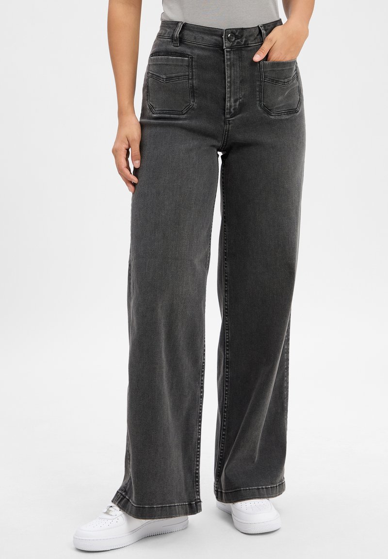 Osoba ubrana w czarne dżinsy o wysokim stanie i szerokich nogawkach z przednimi kieszeniami naszytymi oraz białe trampki, stojąca z jedną ręką w kieszeni.