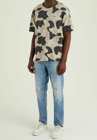 Camiseta de manga corta beige con patrones abstractos en negro, combinada con jeans desgastados de color azul claro y zapatillas blancas.