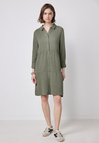 Abito verde oliva con bottoni, colletto a camicia, maniche a tre quarti, design a strati e tessuto testurizzato, che arriva a metà coscia.
