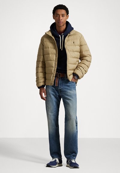Beige quiltet pufferjakke over en marineblå hættetrøje, blå jeans og marineblå sneakers. Har en hætte med snøre og et bælte med farverige striber.