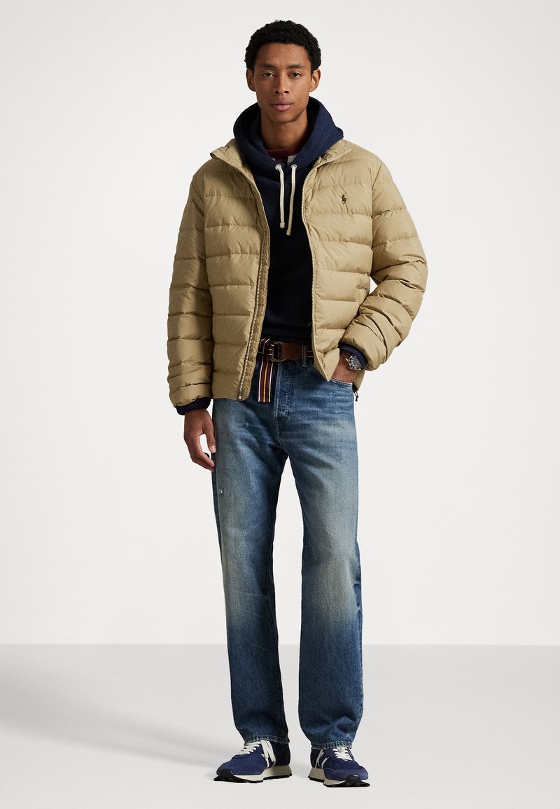 Beige gewatteerde pufferjack over een marineblauwe hoodie, blauwe jeans en marineblauwe sneakers. Heeft een trekkoordcapuchon en een riem met kleurrijke strepen.