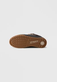 Etnies sporta apavi ar melnu tekstila augšdaļu, brūnu gumijas zoli un teksturētu saķeres rakstu. Raksturo kontrastējošas šņorēšanas un logo detaļas.