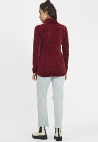 Bordeaux coltrui met een gestructureerde breisel, lange mouwen en een iets langere achterkant. Gecombineerd met lichtblauwe versleten jeans en zwarte boots.