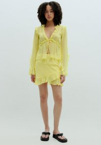 Geel doorschijnende twee-delige outfit met een gerimpelde top met lange mouwen en een strikje aan de voorkant, en een bijpassende gerimpelde mini-jurk. Zwarte sandalen.