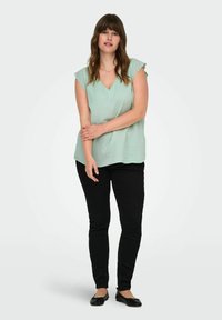 Mintgroene top met geschulpte korte mouwen en een V-hals, gecombineerd met zwarte strakke broek en platte zwarte schoenen. Over het algemeen een gladde textuur.