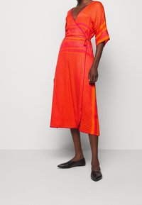 Rutig klänning i orange och rosa med djup V-ringning, korta ärmar, sidoknytning och midi-längd, med en jämn textur och geometrisk design.