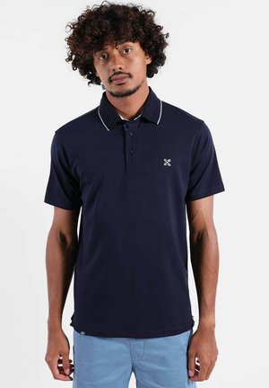 Homme aux cheveux bouclés portant un polo bleu marine avec des bordures bleu clair et un short bleu clair, debout devant un fond blanc uni.
