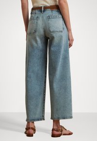 Gaiša džinsa auduma platās kājas bikses ar augstu vidukli, kas iekļauj divas aizmugures kabatas un saplēstu apakšmalu. Saskaņotas ar brūnām siksniņu sandālēm.