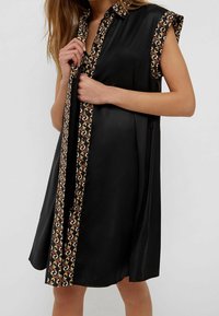 Frau trägt ärmelloses schwarzes Kleid mit gemustertem Besatz entlang der Knopfleiste, am Kragen und an den Armlöchern.