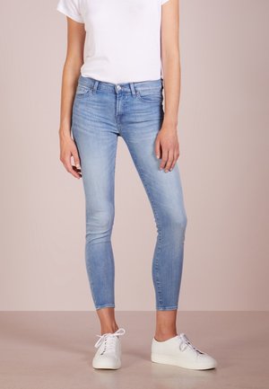 Femme portant un jean skinny bleu clair, un t-shirt blanc uni et des baskets blanches à lacets, debout sur un fond neutre.