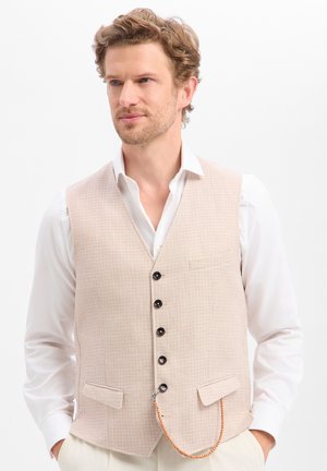 Mann trägt beige strukturierte Weste über weißem Hemd, Hände in den Taschen, schaut leicht nach links vor weißem Hintergrund.