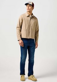 Beige-tyylinen vetoketjullinen takki, jossa on kaulus, yhdistettynä sinisiin farkkuihin ja vaaleanruskeisiin mokkanahkasaappaisiin. Mallilla on myös kaksivärinen lippis, jossa on logo.