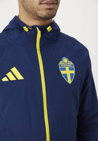 Casaco de marinha com um fecho amarelo, apresentando o logótipo "SVERIGE" e três riscas brancas no lado esquerdo do peito. Fabricado em um material liso e leve.