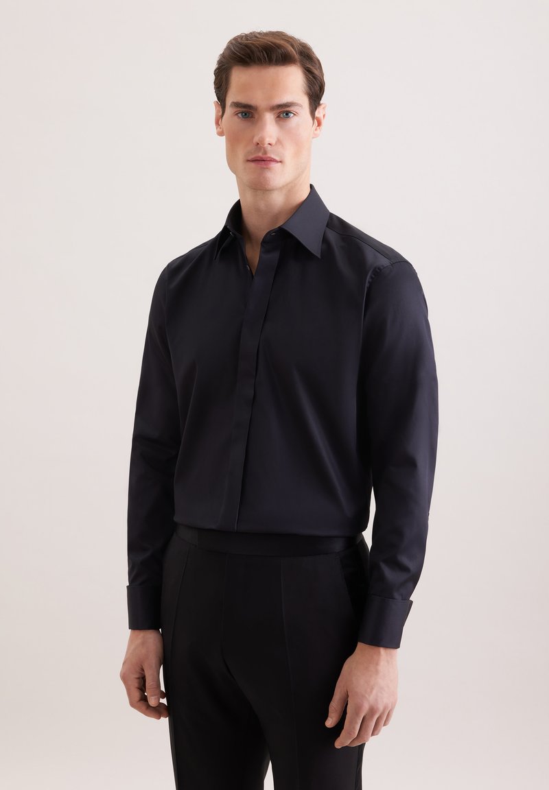 Homme portant une chemise noire ajustée avec des boutons cachés, debout devant un fond clair uni.