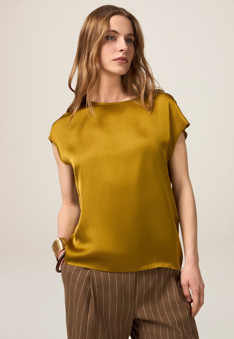 Ärmelloses, senffarbenes Satin-Top mit rundem Ausschnitt und fließender Silhouette, kombiniert mit gestreiften, braunen High-Waist-Hosen.