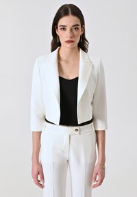 Veste courte blanche en tissu lisse, avec un col classique, des manches trois-quarts et une coupe structurée, portée sur un haut noir.