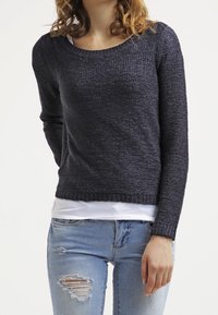 Granatowy sweter dziany z szerokim dekoltem, długimi rękawami i teksturowanym wykończeniem. Nosi się go na białą koszulę, w zestawieniu z uszkodzonymi jasnoniebieskimi dżinsami.