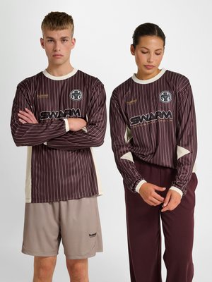 Twee modellen dragen bijpassende bordeauxrode en beige gestreepte sportshirts met lange mouwen en logo's, de ene gecombineerd met beige short, de andere met bordeauxrode broek.