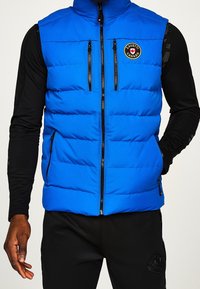 Blaues isoliertes Weste mit hohem Kragen, Frontreißverschluss und mehreren Reißverschlusstaschen. Verfügt über gepolsterten Textur und ein rundes Logo-Patch.