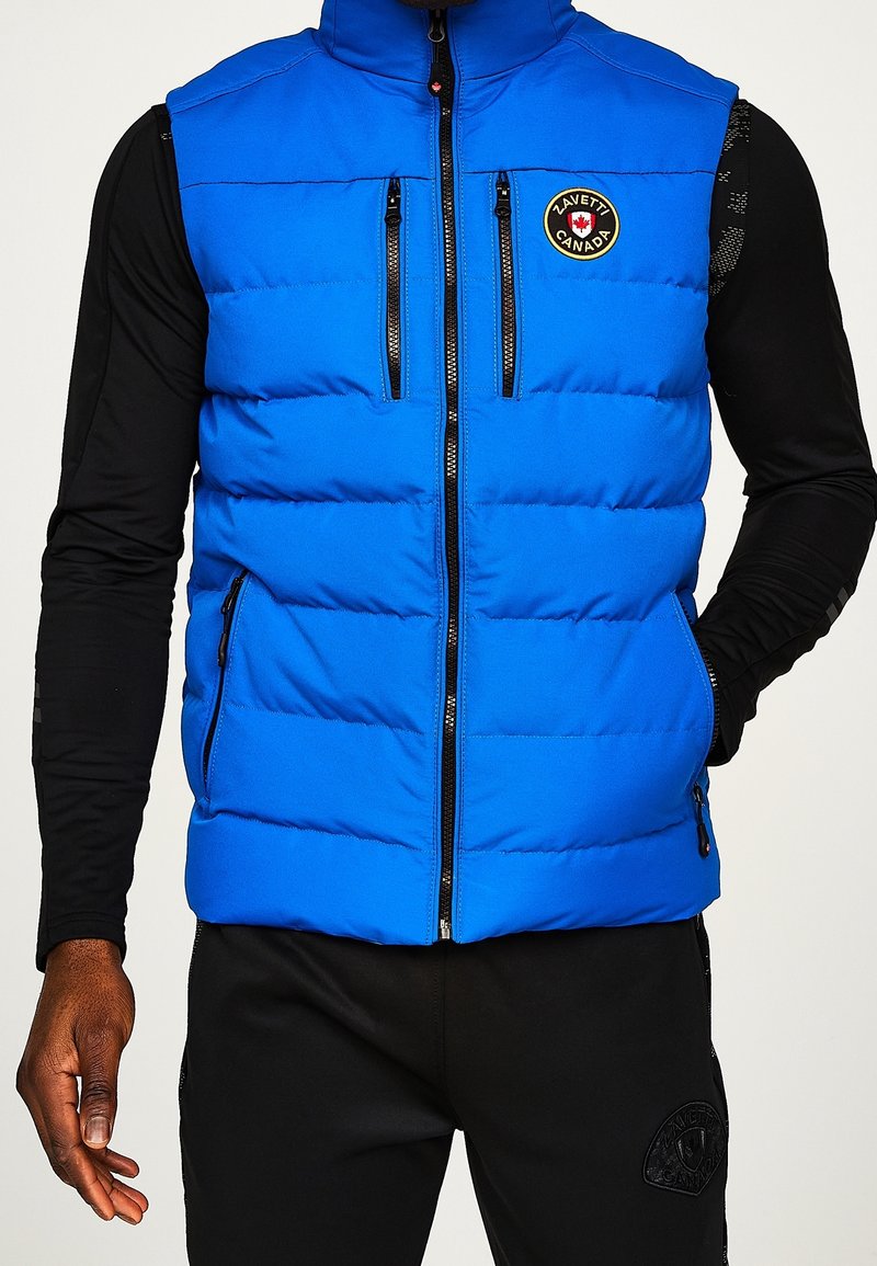 Blaues isoliertes Weste mit hohem Kragen, Frontreißverschluss und mehreren Reißverschlusstaschen. Verfügt über gepolsterten Textur und ein rundes Logo-Patch.