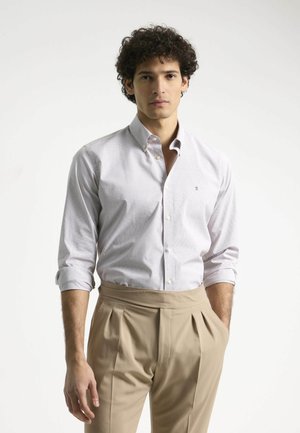 Camisa blanca de botón con un sutil patrón de cuadrícula, mangas enrolladas, combinada con pantalones beige de cintura alta. Logo simple en la camisa.