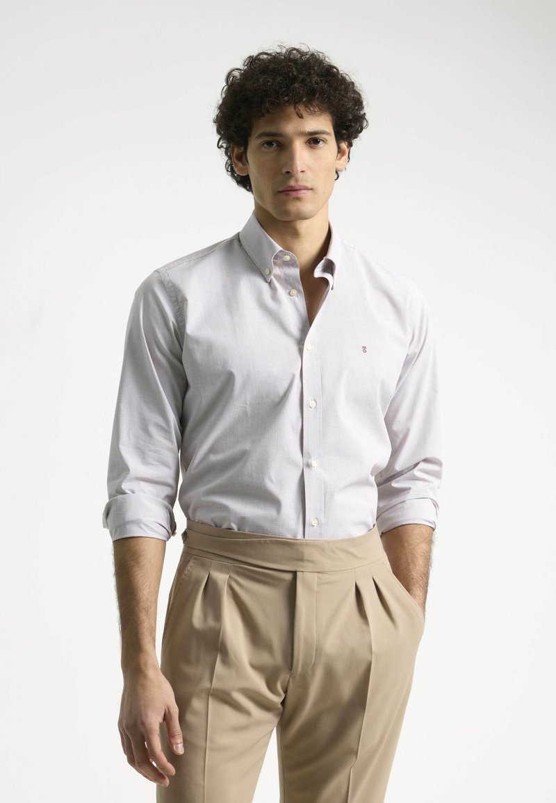 Camisa blanca de botón con un sutil patrón de cuadrícula, mangas enrolladas, combinada con pantalones beige de cintura alta. Logo simple en la camisa.