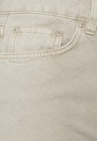 Tejido de denim color beige claro que presenta un bolsillo frontal con remaches de metal plateado y detalles de costura contrastantes. Textura suave con un ligero estiramiento.