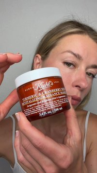 Mujer sosteniendo cerca de su rostro un frasco de la mascarilla revitalizante de luminosidad de cúrcuma y semillas de arándano de Kiehl's.