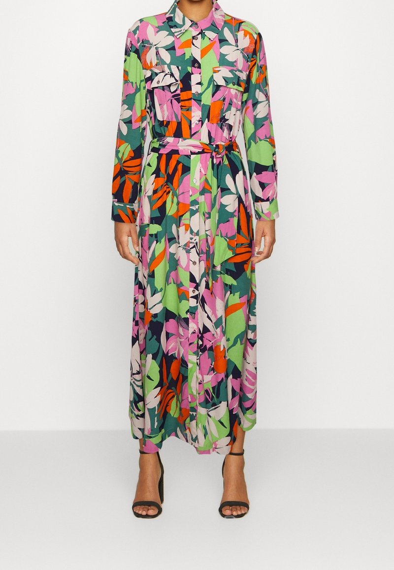 Femme portant une robe fleurie à manches longues, boutonnée, avec des motifs de feuilles vertes, roses, orange et bleu marine, ceinturée à la taille, associée à des talons noirs.